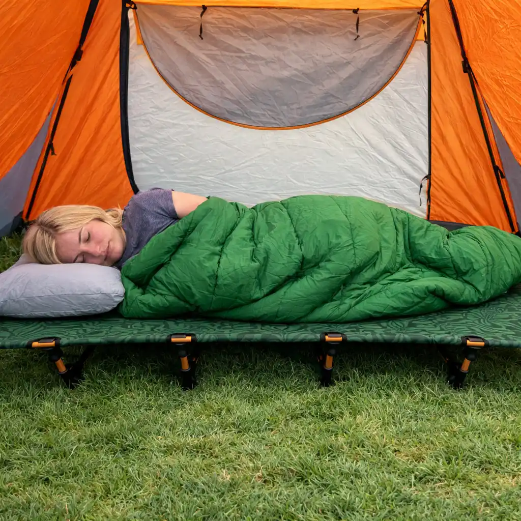 Blonde woman sleeping on a camping cot inside an orange tent in a green sleeping bag