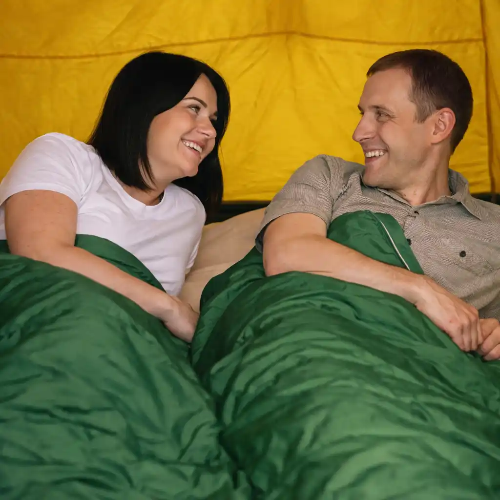 Couple relaxing inside a bright yellow tent in green sleeping bags, smiling at each other.