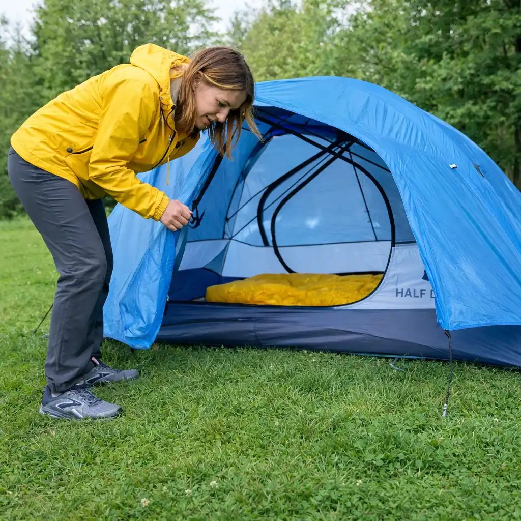Woman in a yellow jacket opening a blue camping tent set up on green grass in a forest.