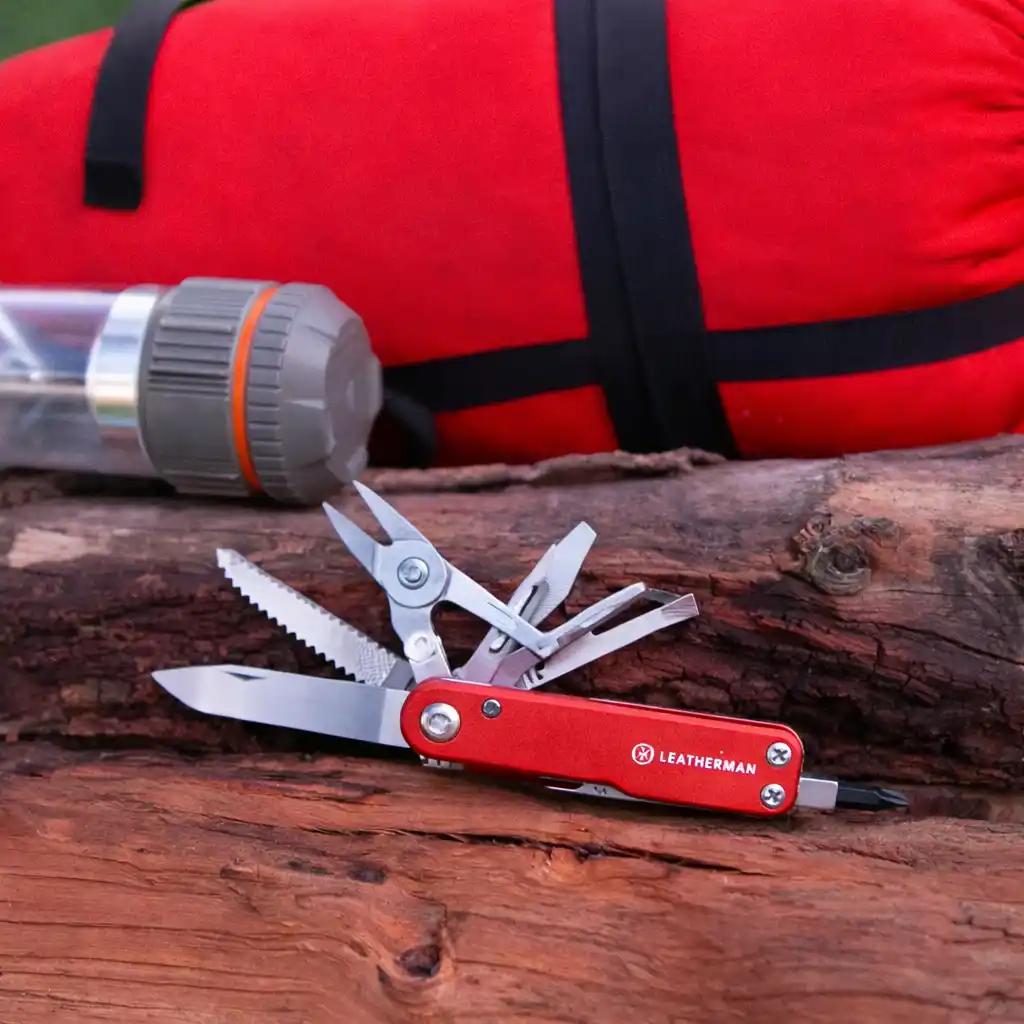 A red-handled multi-tool resting on a piece of tree bark in a natural setting, with a vivid red travel bag and camping gear behind it