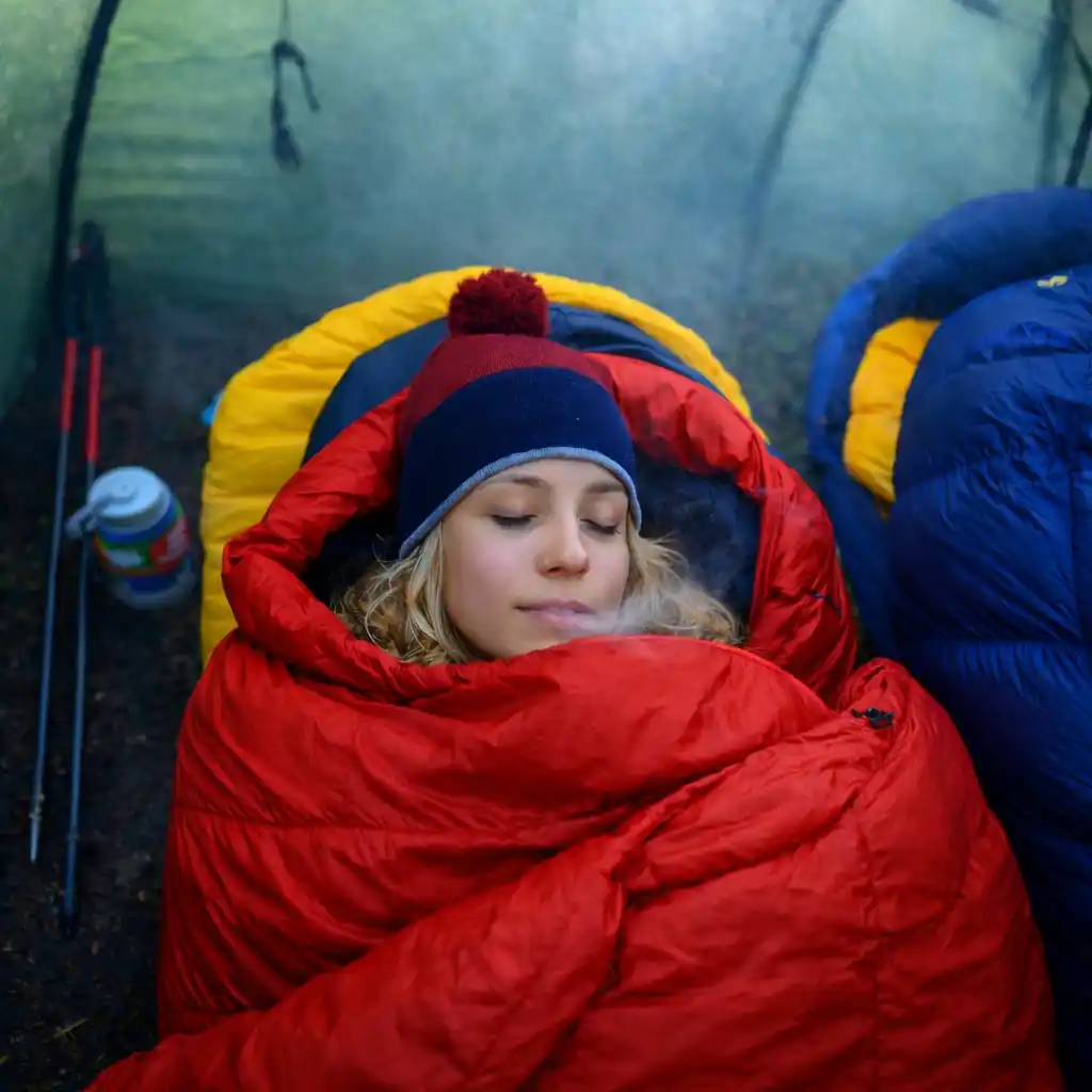 Blonde woman sleeping inside a tent in a red sleeping bag, faint visible breath in cool humid air, illustrating heat loss and moisture buildup overnight