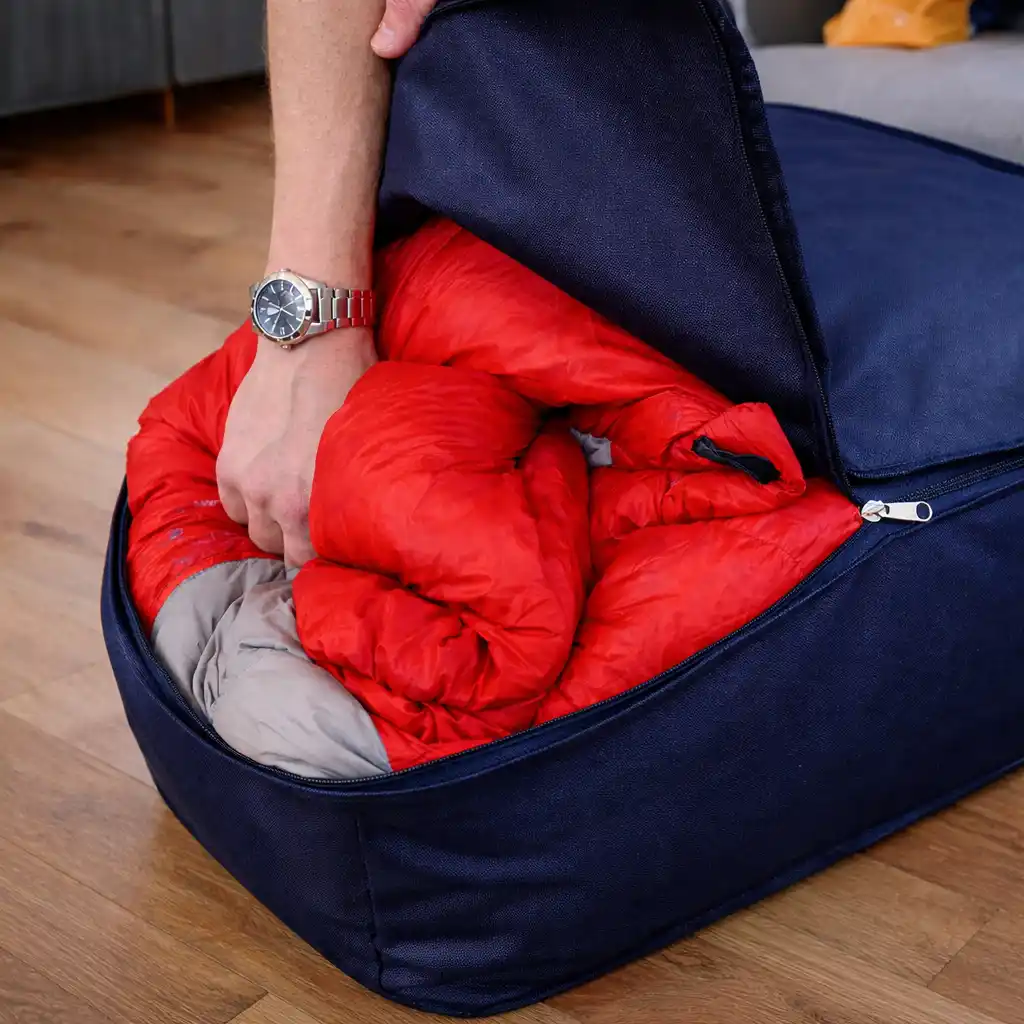 Red sleeping bag being stored in a dark blue storage bag indoors, showing proper loose storage to prevent insulation compression and loss of warmth over time.
