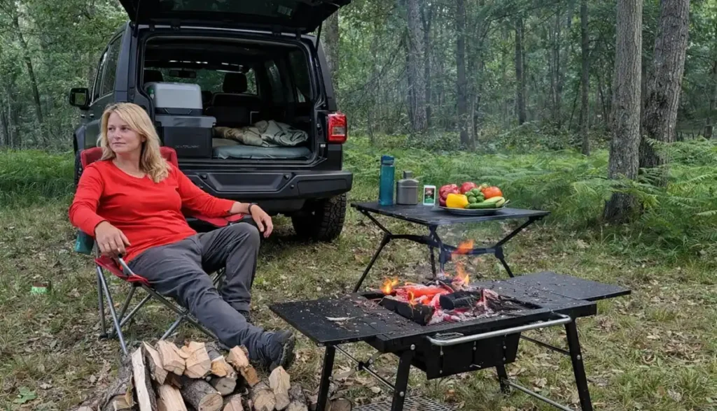 Blonde woman in a red shirt relaxing at a forest campsite next to a Jeep, with a campfire and fresh vegetables on a table