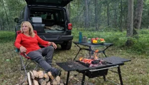 Blonde woman in a red shirt relaxing at a forest campsite next to a Jeep, with a campfire and fresh vegetables on a table