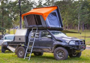 Silver pickup truck with orange rooftop tent parked by a mountain lake surrounded by pine forest