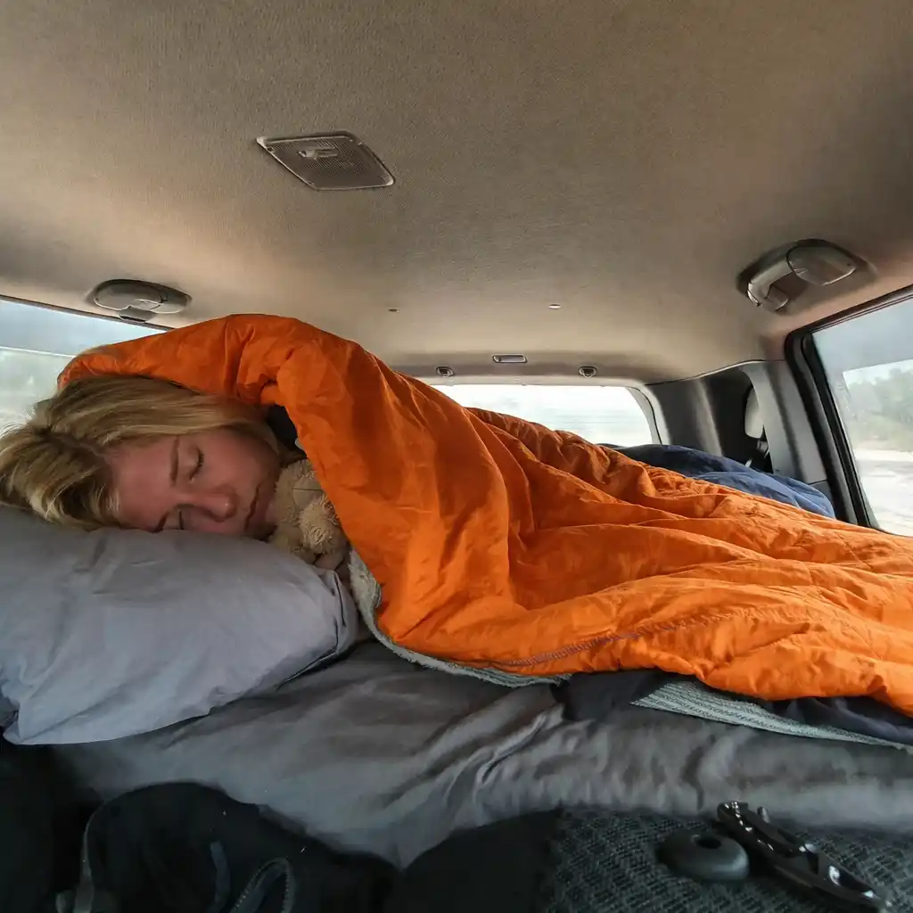 sleeping inside car camping setup with mattress pillow and orange sleeping bag in vehicle interior