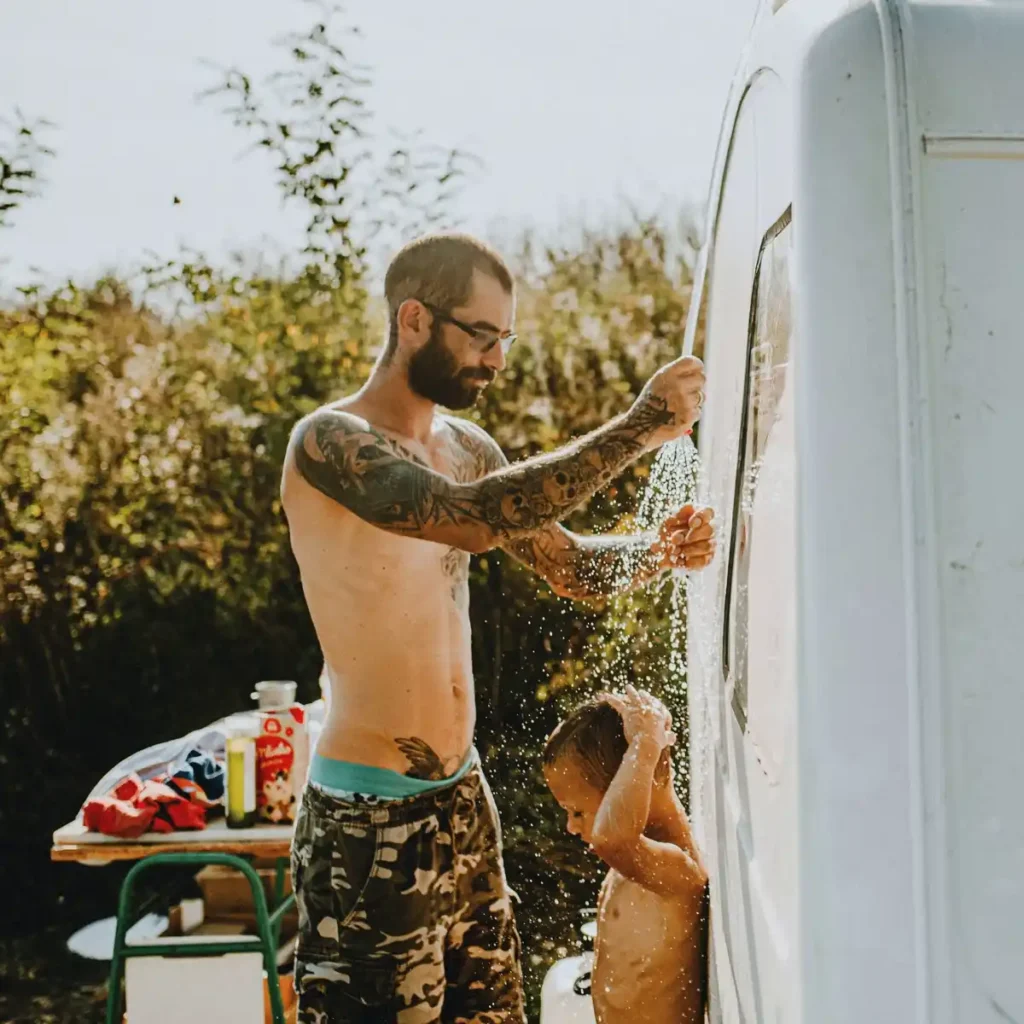 A tattooed father helping his young son take an outdoor shower next to a white camper van on a sunny day.